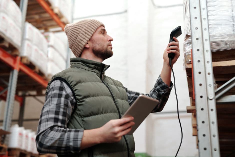 A warehouse worker scanning a product barcode next to shelves of inventory, checking listing and stock details
