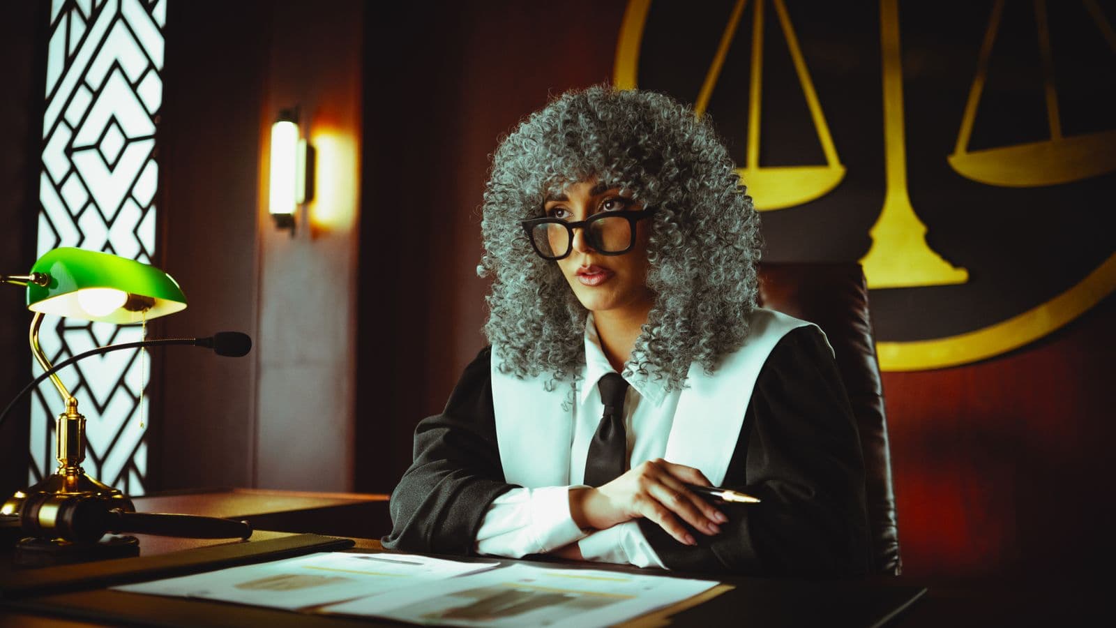 A judge in courtroom robes seated at a desk with the scales of justice in the background.